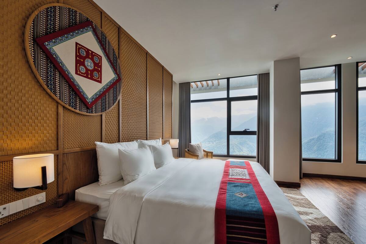Ein Hotelzimmer mit einem großen Bett, einer Wanddekoration, einem Nachttisch mit Lampe und Blick auf Berge durch große Fenster.