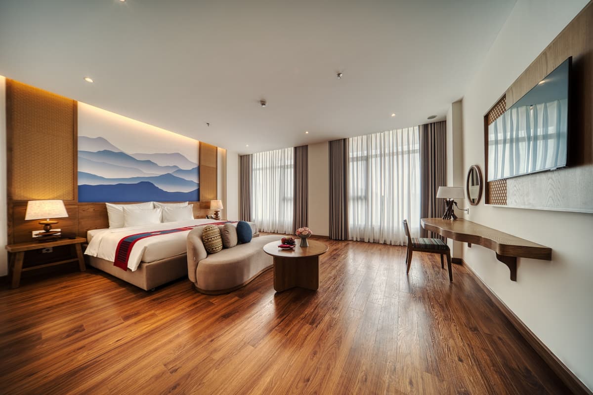 Ein Hotelzimmer mit Bett, Sofa, kleinem runden Tisch, Schreibtisch und großem Fenster mit Vorhängen.