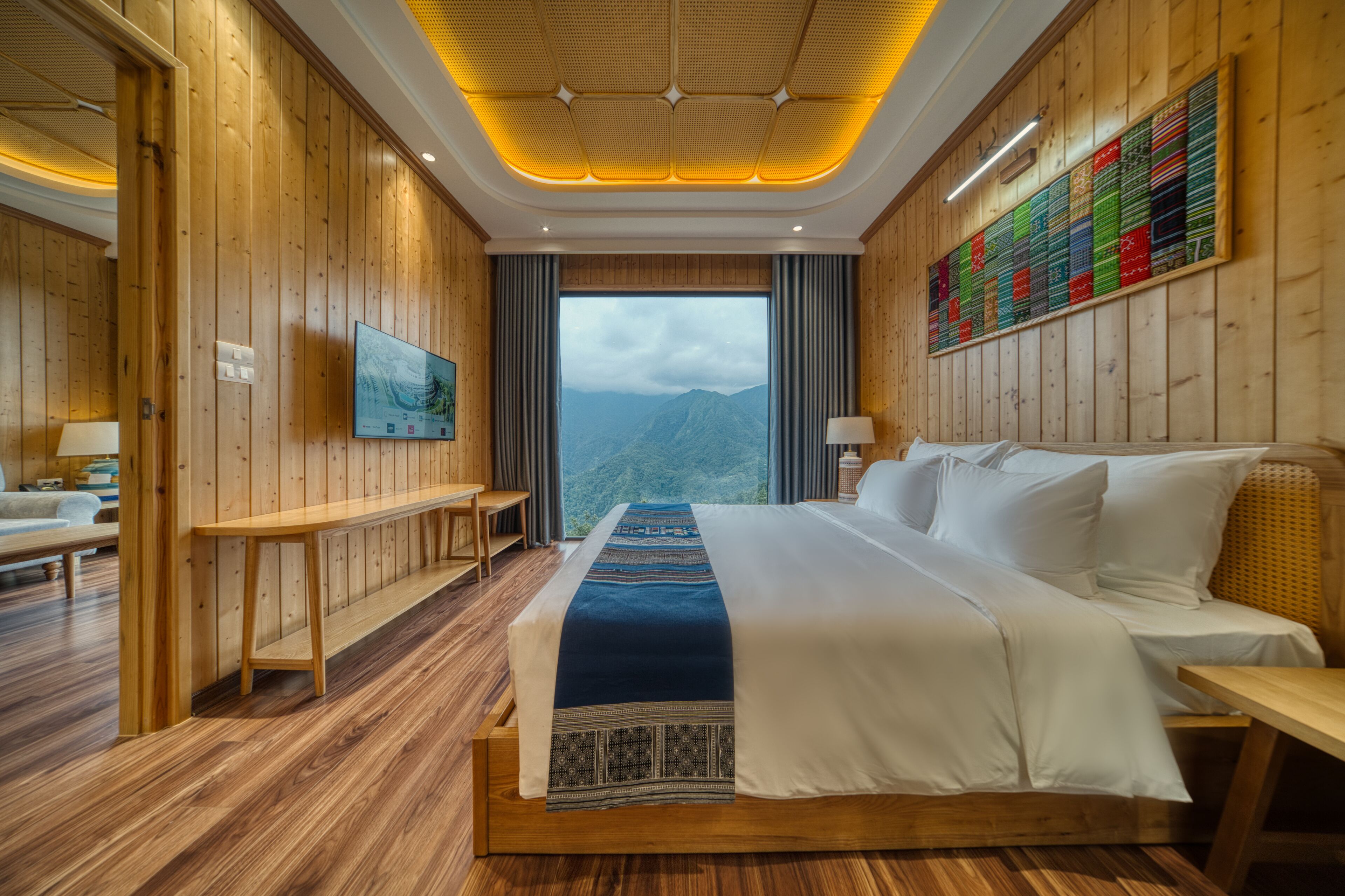 A hotel room with a large bed, wooden walls, a view of mountains, a long table, and a wall art piece.