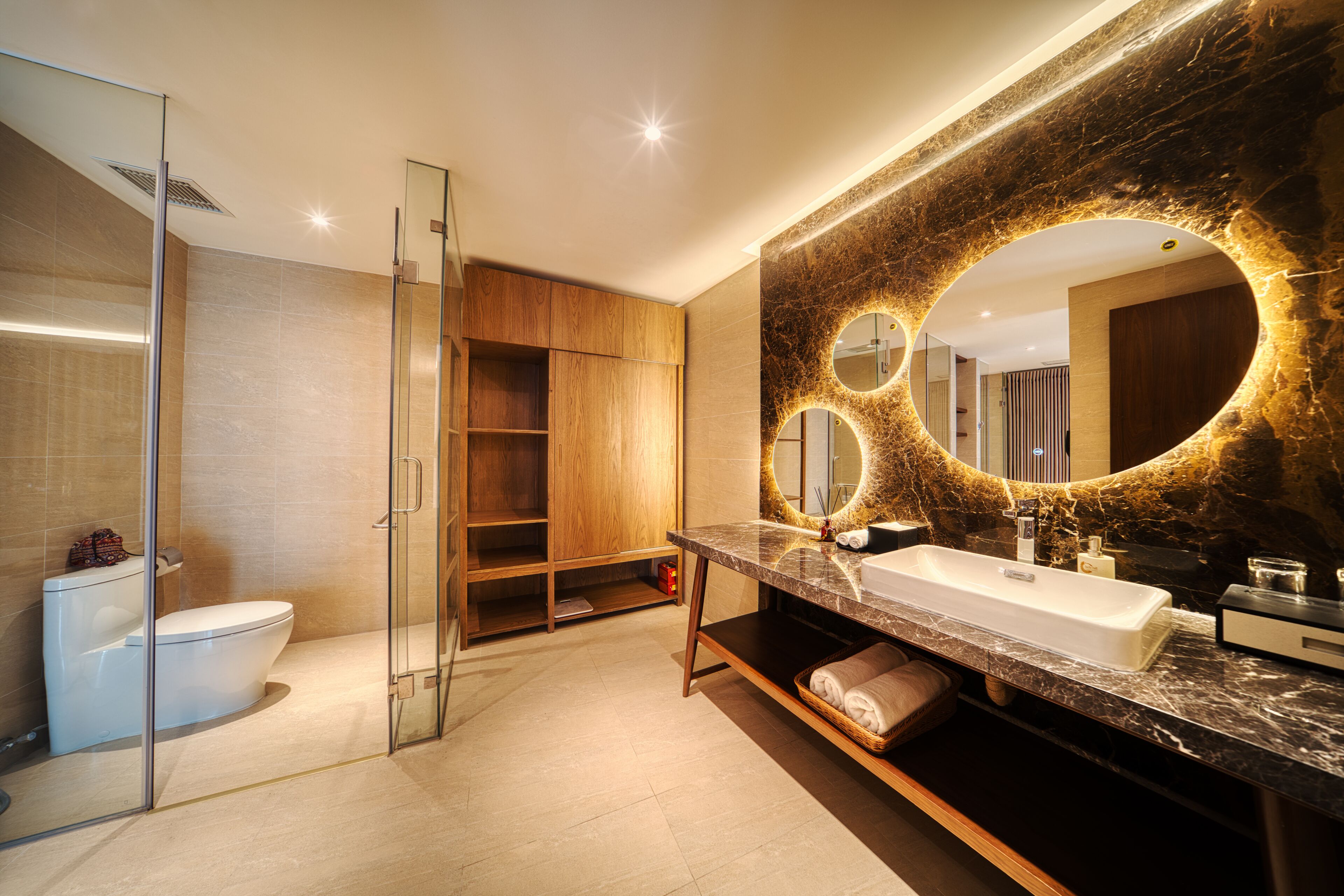 Un baño moderno con ducha de vidrio, tocador grande con encimera de mármol y espejo redondo con marco dorado.