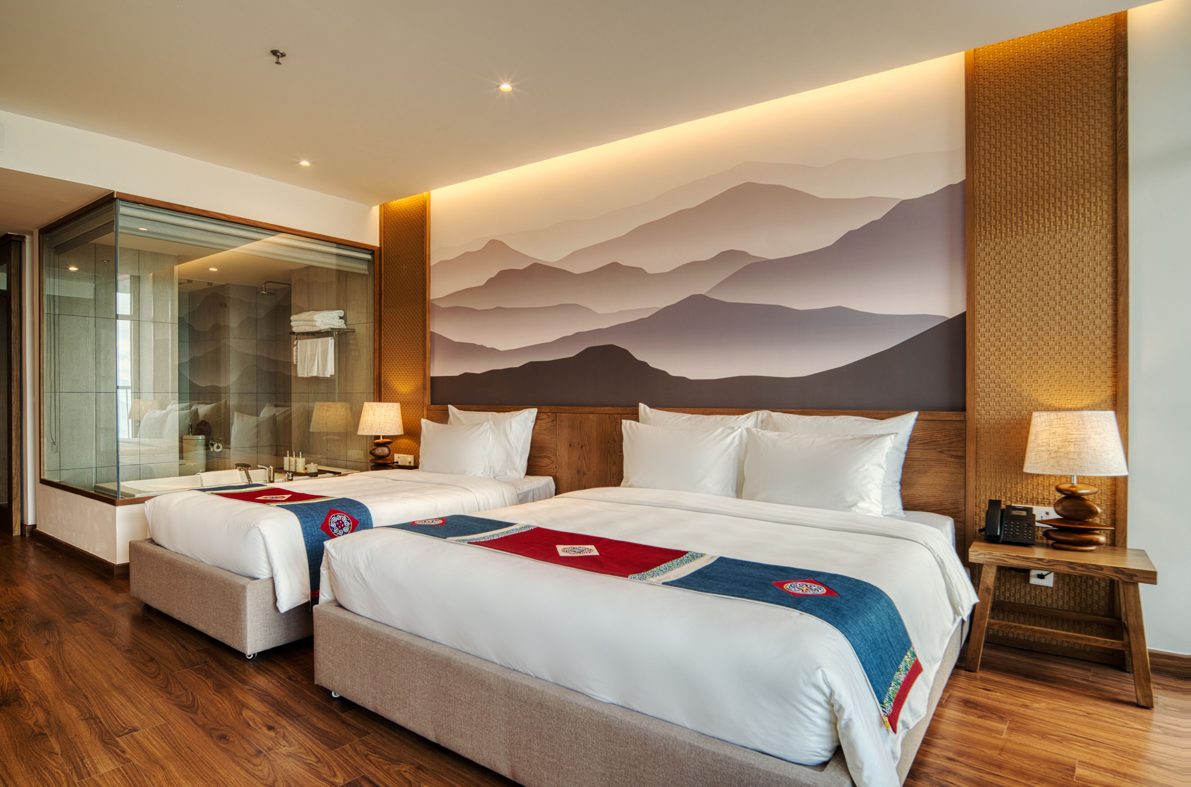 A hotel room with two beds, a glass shower enclosure, a wooden headboard, and a mountain mural on the wall.