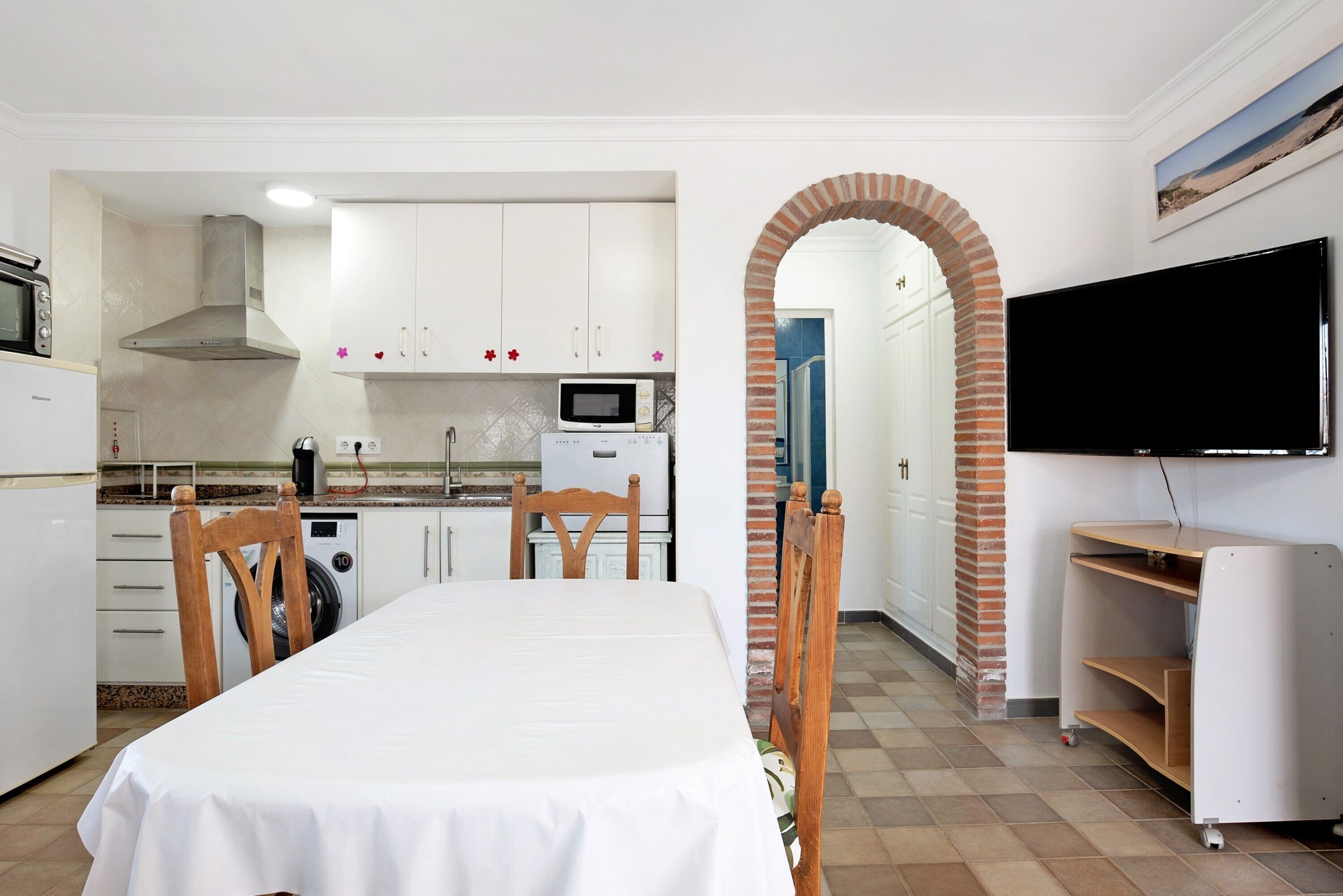 Une cuisine avec des meubles blancs, un four à micro-ondes et une cuisinière. On y trouve une table à manger avec des chaises. Un arc donne accès à une autre pièce où se trouve un téléviseur.