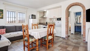 A compact kitchen with white cabinets, a dining table with chairs, a sofa, and a large window.