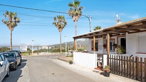 A street with parked cars, a white building with a wooden fence, and palm trees.