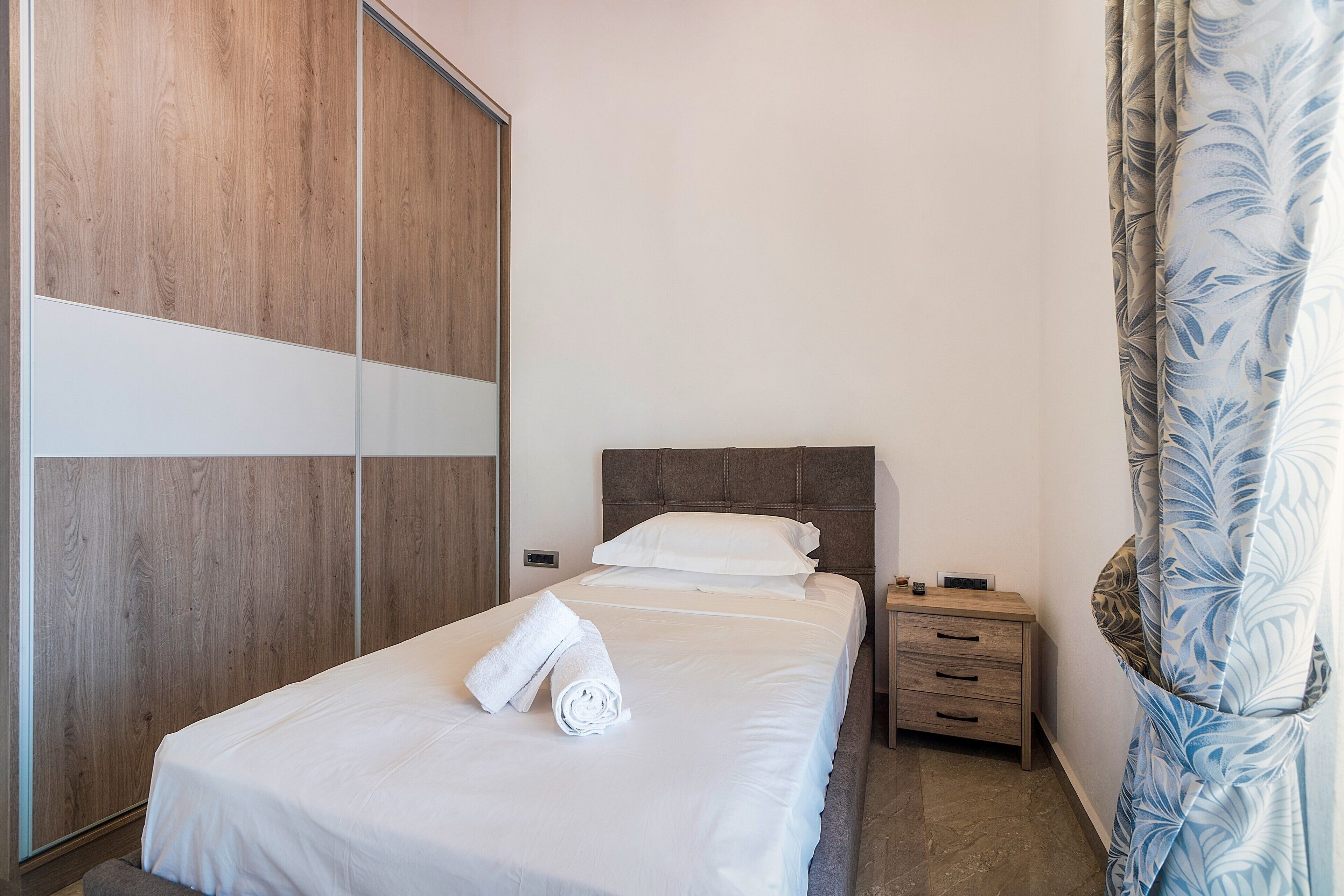 Een net opgemaakt bed met een witte dekbedovertrek en opgerolde handdoeken, een houten garderobe, een nachtkastje met een lamp en een bloemenprint gordijn.