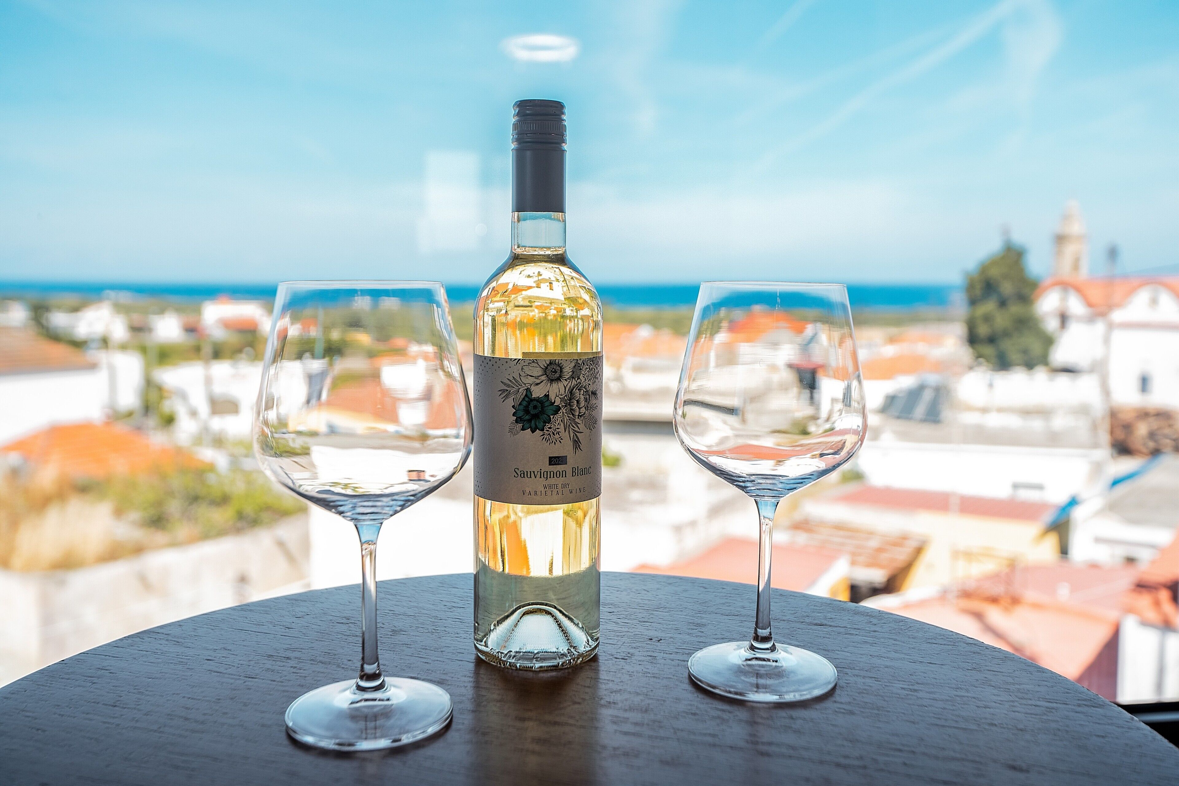Una botella de Sauvignon Blanc y dos copas sobre una mesa con vista a edificios y al mar.