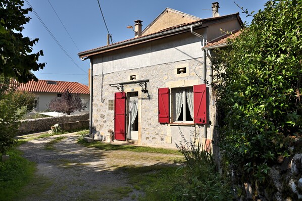 Un bâtiment en pierre avec des volets rouges, un chemin de gravier et un mur en pierre.