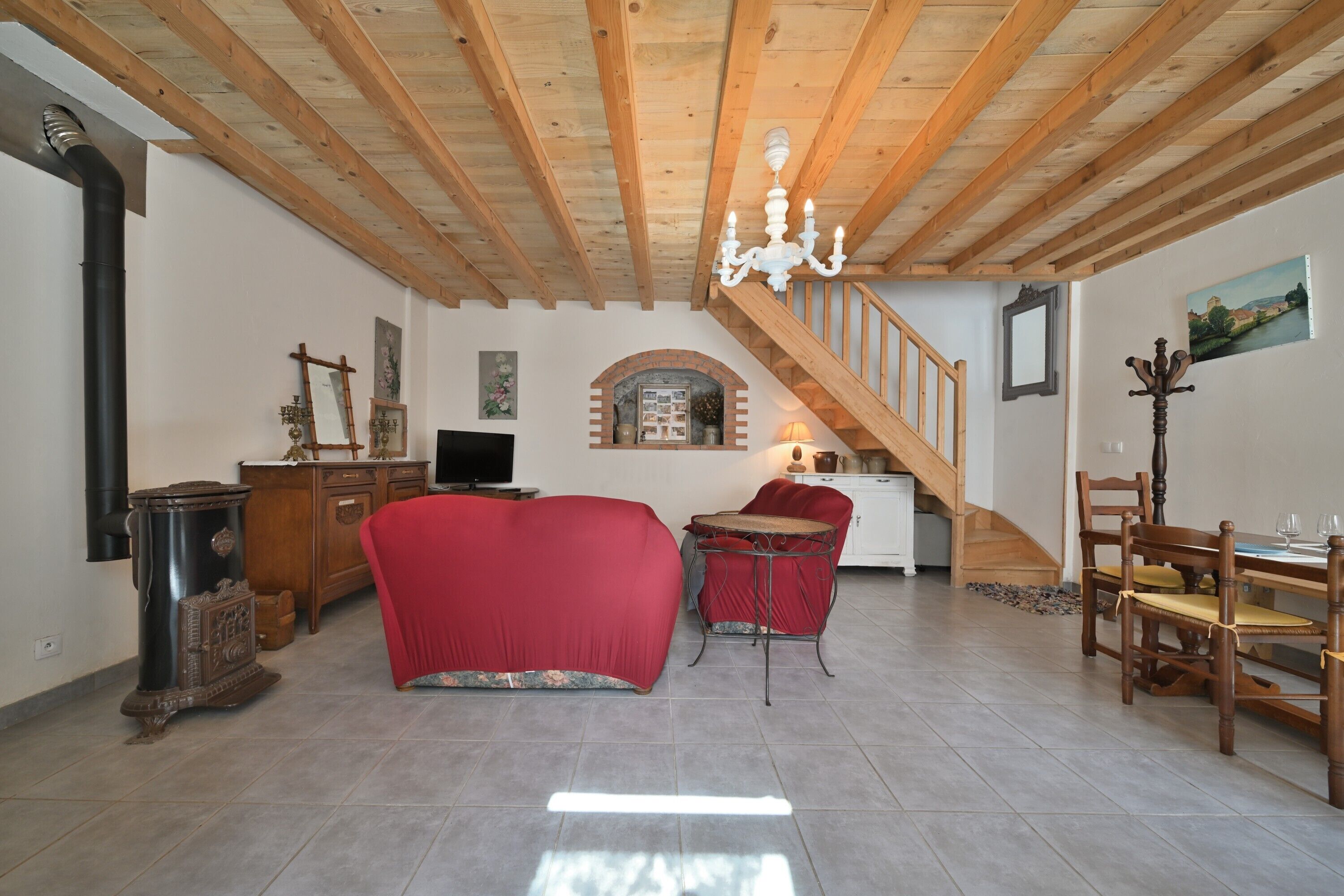 A spacious living area with a wooden ceiling, a staircase, a dining table with chairs, and a red sofa.