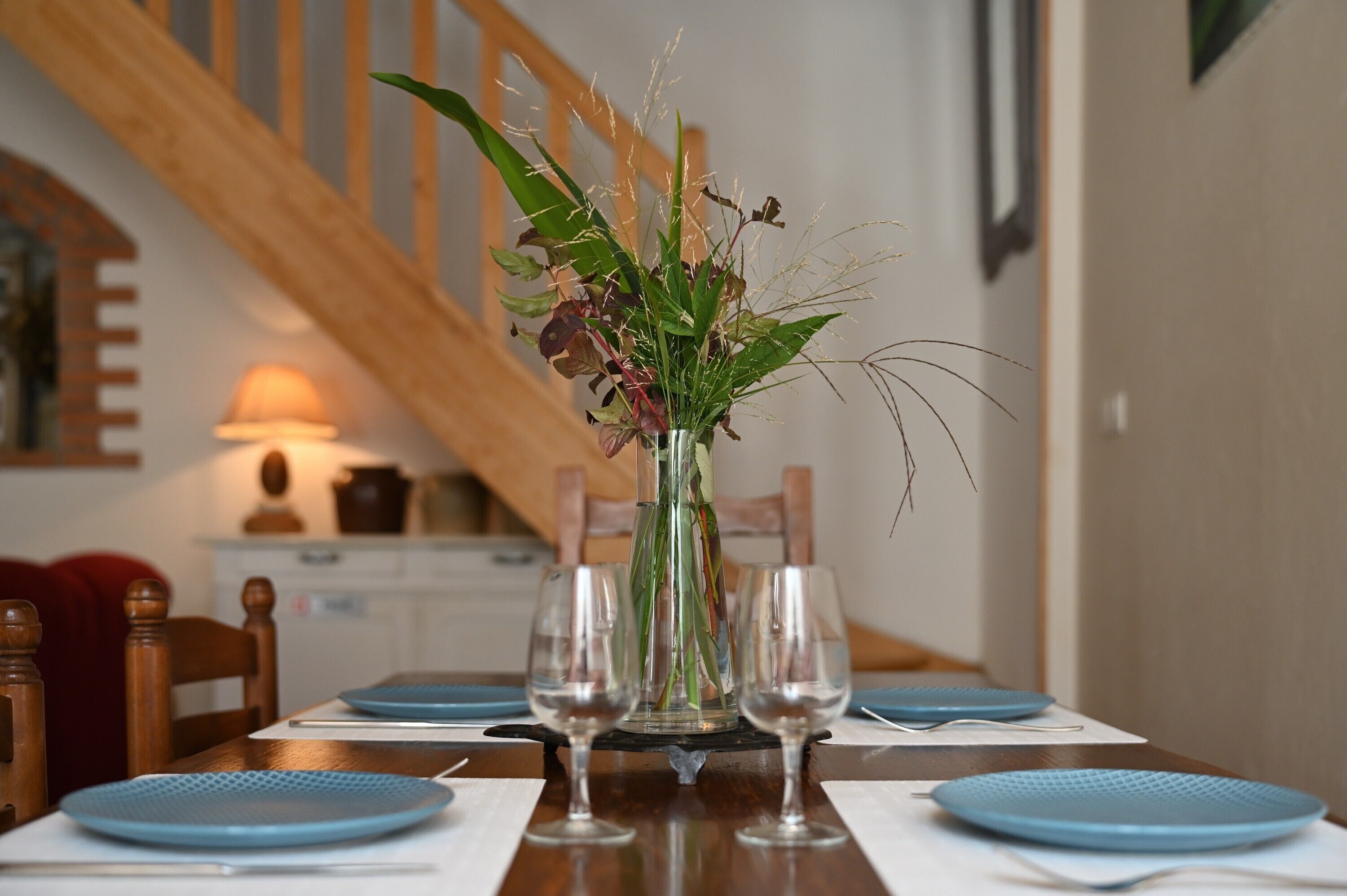 A dining table with blue placemats, clear glassware, and a vase with green and purple foliage.