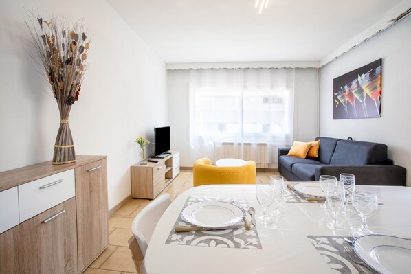 Ein modernes Wohnzimmer mit einer Couch, einem Esstisch gedeckt mit Tellern und Gläsern, einem Fernseher und einem Fenster mit Vorhängen.