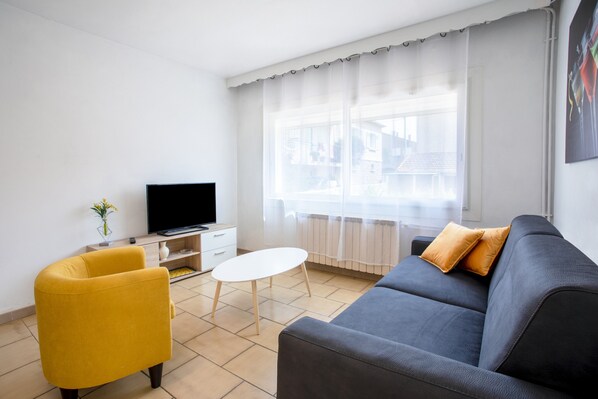 Un salon moderne avec un canapé, un fauteuil jaune, une petite table blanche, un téléviseur et une fenêtre ornée de rideaux légers.