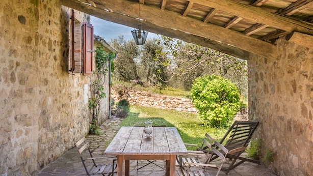 A stone patio with a wooden table and chairs, a hanging lantern, and a view of a garden and stone wall.