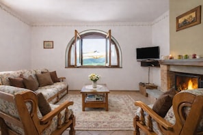 Un salon comprenant un canapé, des fauteuils, une cheminée, un téléviseur et une grande fenêtre surmontée d’une arche.