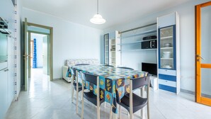 A dining area with a table, chairs, a hanging light fixture, and a kitchen area with shelves and appliances. - Apartment "Residenza del Sole 1" with Shared Pool & A/C (Villasimius)