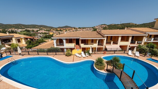 A swimming pool with a deck, surrounded by buildings with balconies and a view of a hilly landscape. - Apartment "Residenza del Sole 1" with Shared Pool & A/C (Villasimius)