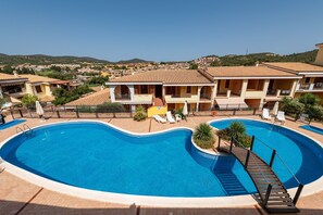 A swimming pool with a deck, surrounded by buildings with balconies and a view of a hilly landscape.