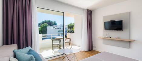 A modern hotel room with a sofa, a bed, a TV, a balcony with a table and chairs, and a view of the city.