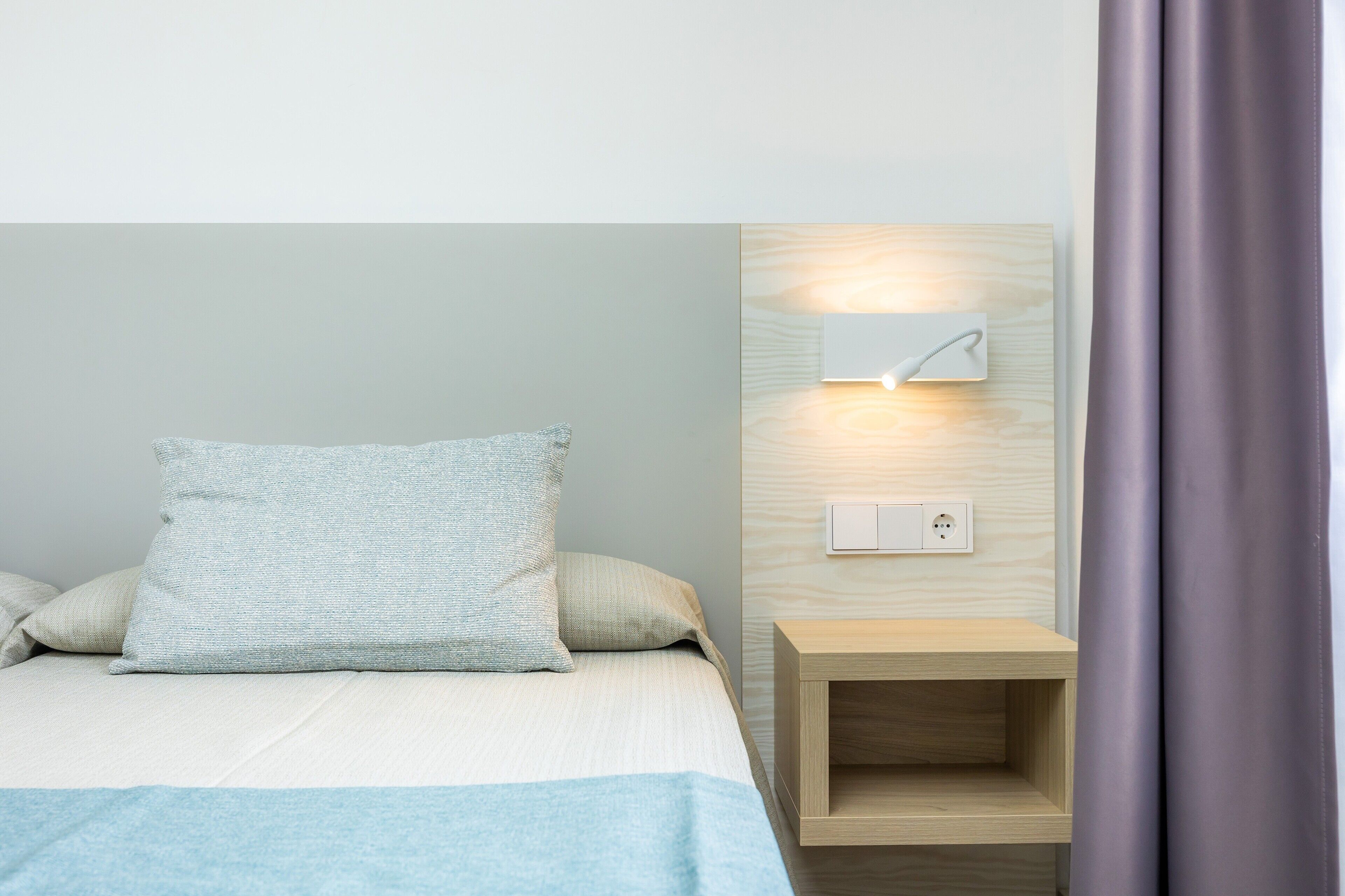 Un lit avec une couverture bleu clair, un oreiller gris et une table de chevet en bois sur laquelle est fixée une lampe murale.