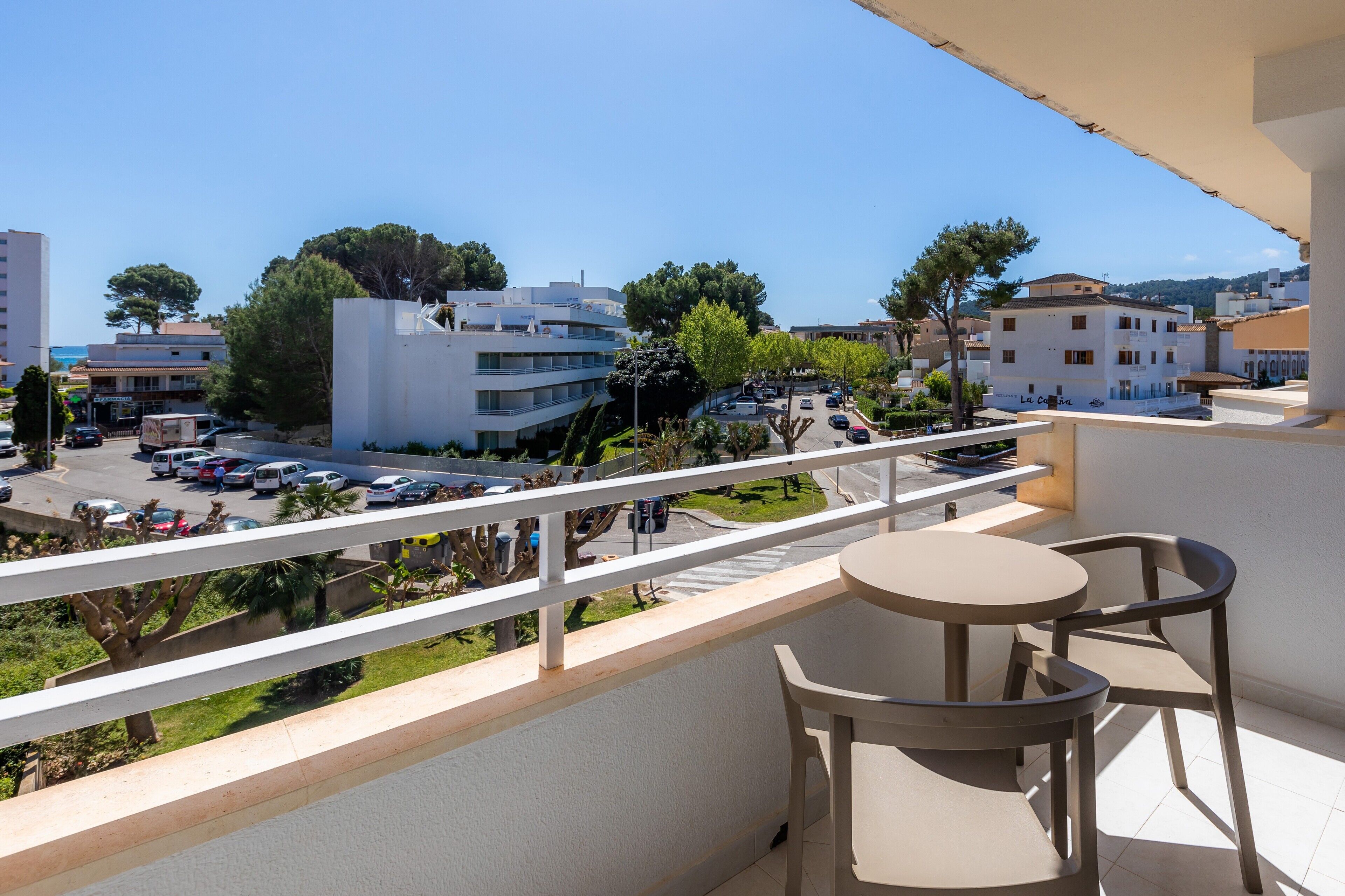 A balcony with a table and chairs, overlooking a street with parked cars and buildings.