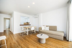 En moderne stue med trægulv, en beige sofa, et rundt sofabord og et spiseområde med hvide stole og et træbord.