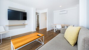 Een moderne woonkamer met een flatscreen televisie, een houten salontafel, een grijze bank met een geel kussen, en een eethoek met witte stoelen en een houten tafel.