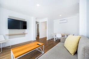A modern living room with a flat-screen TV, a wooden coffee table, a gray sofa with a yellow pillow, and a dining area with white chairs and a wooden table.
