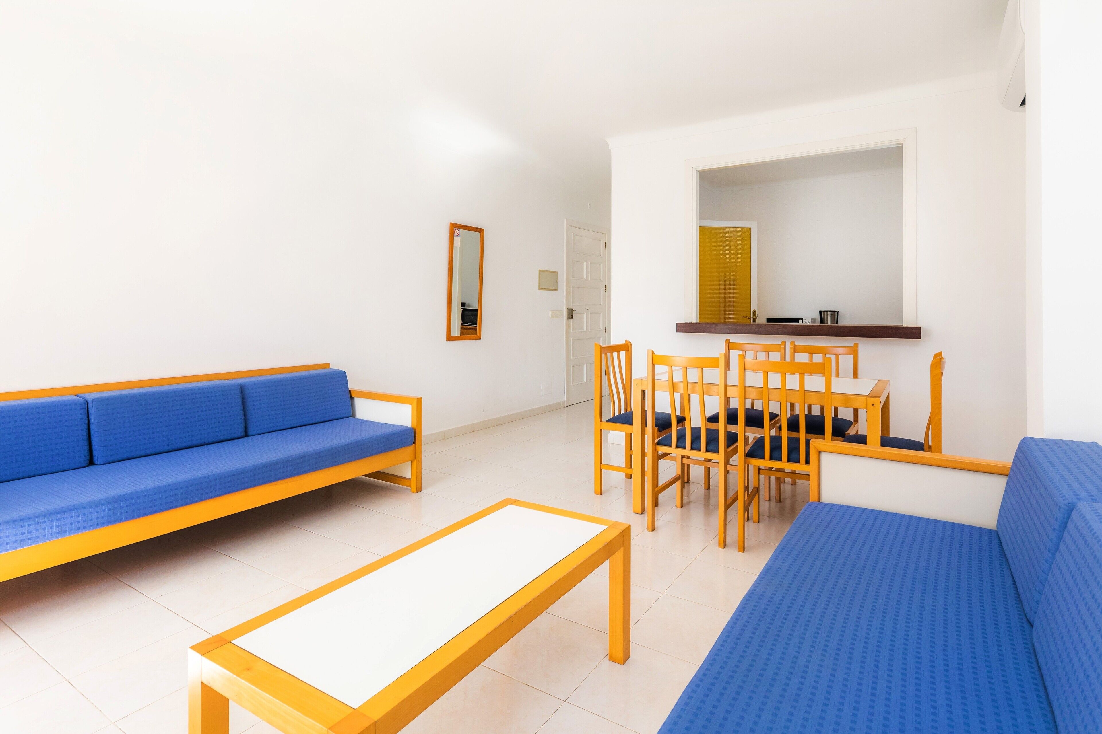 Una habitación con un sofá azul, una mesa de comedor de madera con sillas, una mesita blanca y un espejo en la pared.