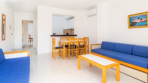 Una sala de estar con un sofá azul, una mesa de comedor de madera con sillas, y una cocina visible a través de un vano abierto.