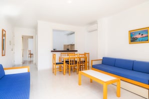 Una sala de estar con un sofá azul, una mesa de comedor de madera con sillas, y una cocina visible a través de un vano abierto.