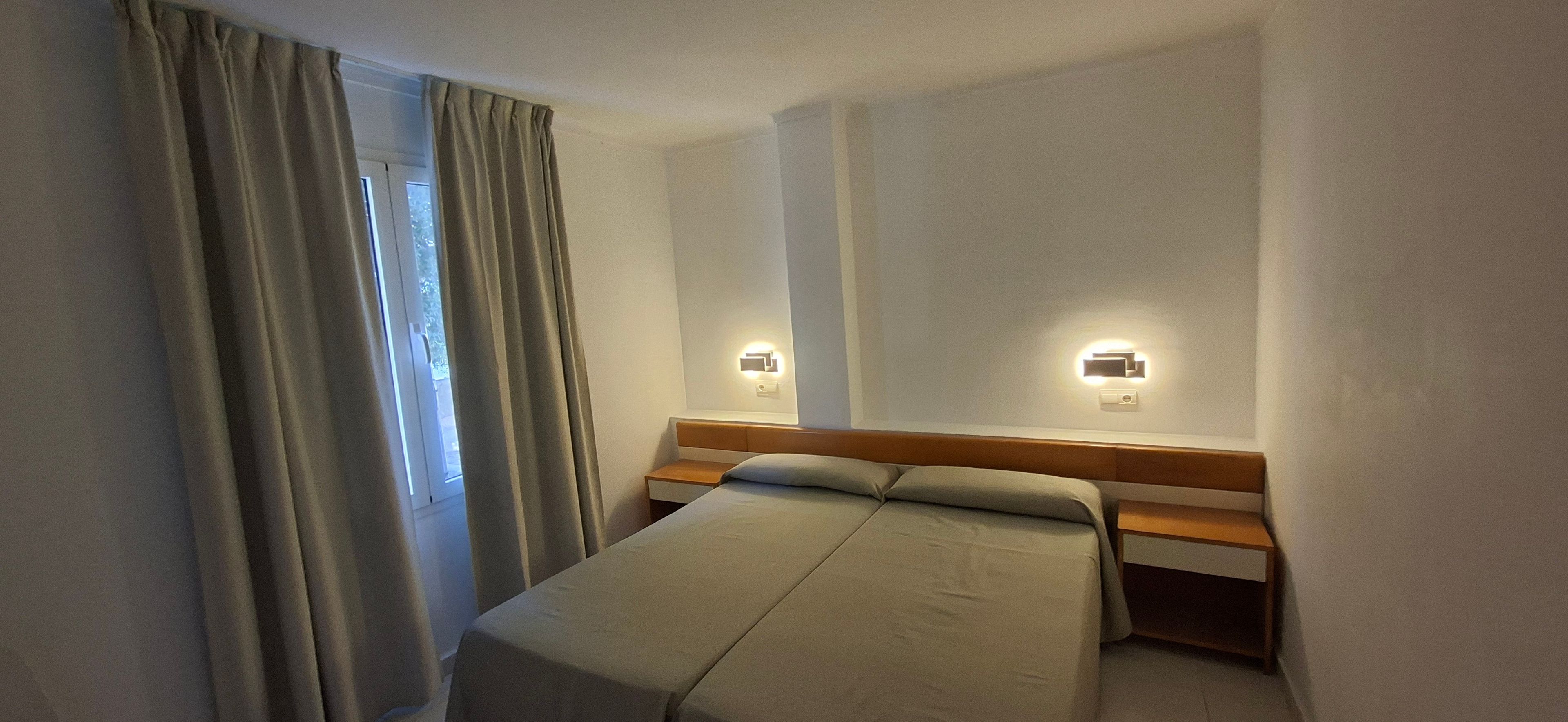 Un lit simple avec une tête de lit en bois, deux appliques murales et une fenêtre avec des rideaux.