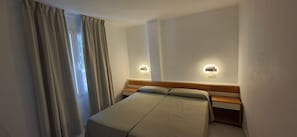 A single bed with a wooden headboard, two wall-mounted light fixtures, and a window with curtains.