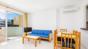 Ein modernes Wohnzimmer mit einem blauen Sofa, einem Holzesstisch und Stühlen, einem Fernseher und einer Schiebeglasdoor zum Balkon.