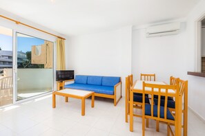 Een moderne woonkamer met een blauwe bank, een houten eettafel en stoelen, een televisie, en een schuifpui naar een balkon.