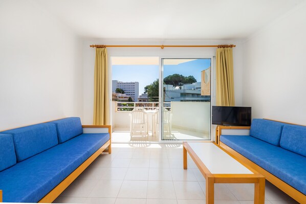 A room with two blue sofas, a wooden table, a TV, and a sliding glass door leading to an outdoor balcony.