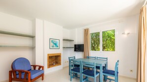 Una habitación con una silla de madera, una mesa azul con cuatro sillas, un televisor, una chimenea y una ventana con cortinas.