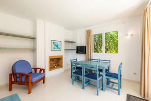 A room with a wooden chair, a blue table with four chairs, a TV, a fireplace, and a window with curtains.