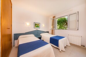 A room with two beds, a wooden wardrobe, a radiator, and a window with a view of greenery.