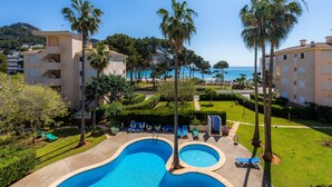 A swimming pool with two separate sections, surrounded by palm trees and lounge chairs, with a view of the sea and buildings in the background. - Apartment 'Oro del Mar IV 05' with Shared Pool, Shared Garden and Wi-Fi (Canyamel)