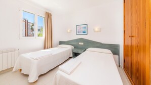 A hotel room with two single beds, a wooden wardrobe, a window with curtains, a wall-mounted picture, and a bedside table with a lamp. - Apartment 'Oro del Mar IV 05' with Shared Pool, Shared Garden and Wi-Fi (Canyamel)