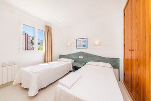 A hotel room with two single beds, a wooden wardrobe, a window with curtains, a wall-mounted picture, and a bedside table with a lamp.
