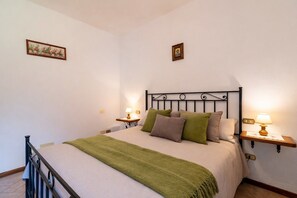 Una cama bien hecha con una manta verde, dos mesitas de noche con lámparas y un cuadro pequeño en la pared.