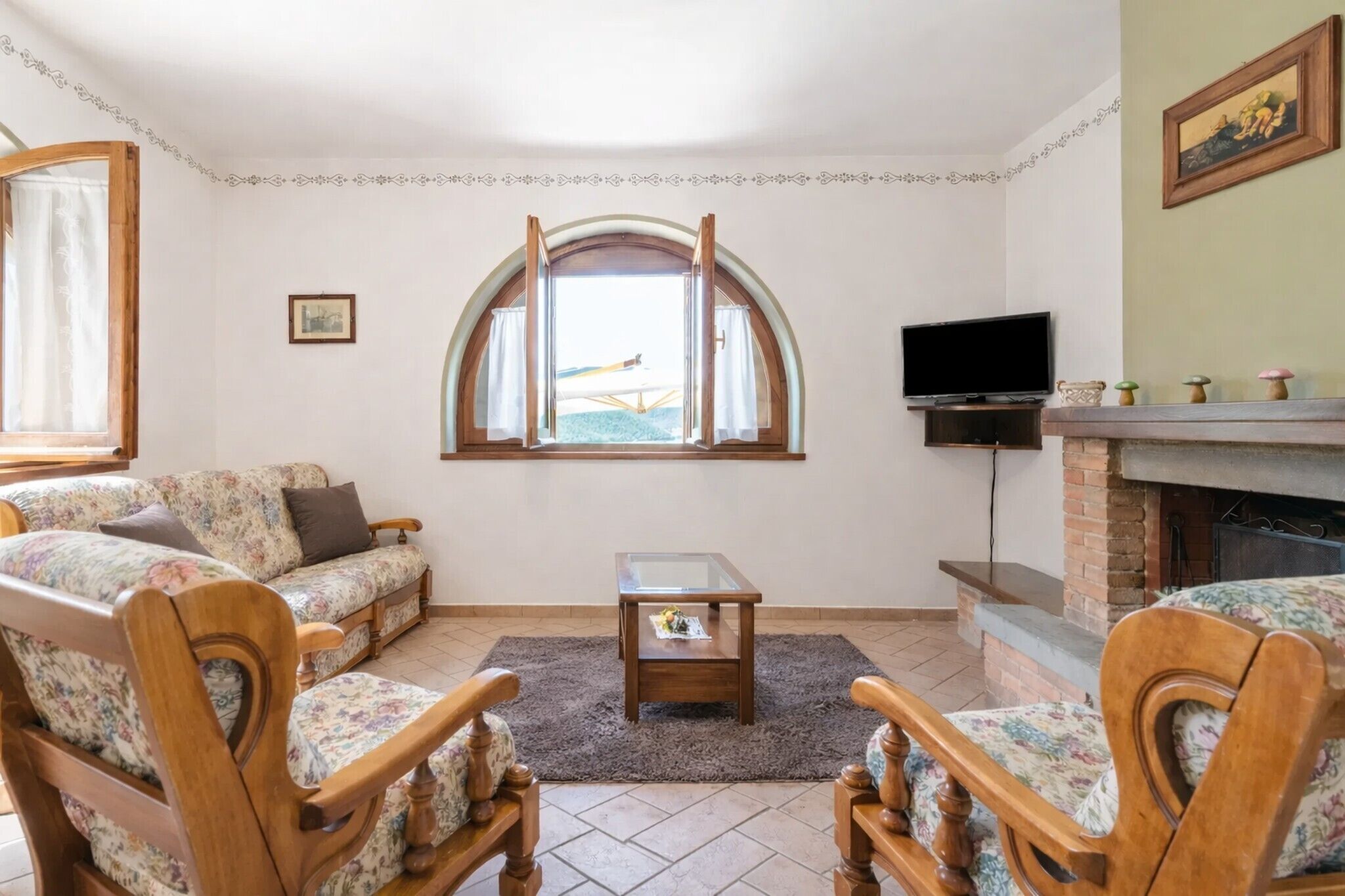 Ein Wohnzimmer mit einer Couch, Sesseln, einem Glastisch, einem Kamin, einem Fernseher und einem Fenster mit Vorhängen.