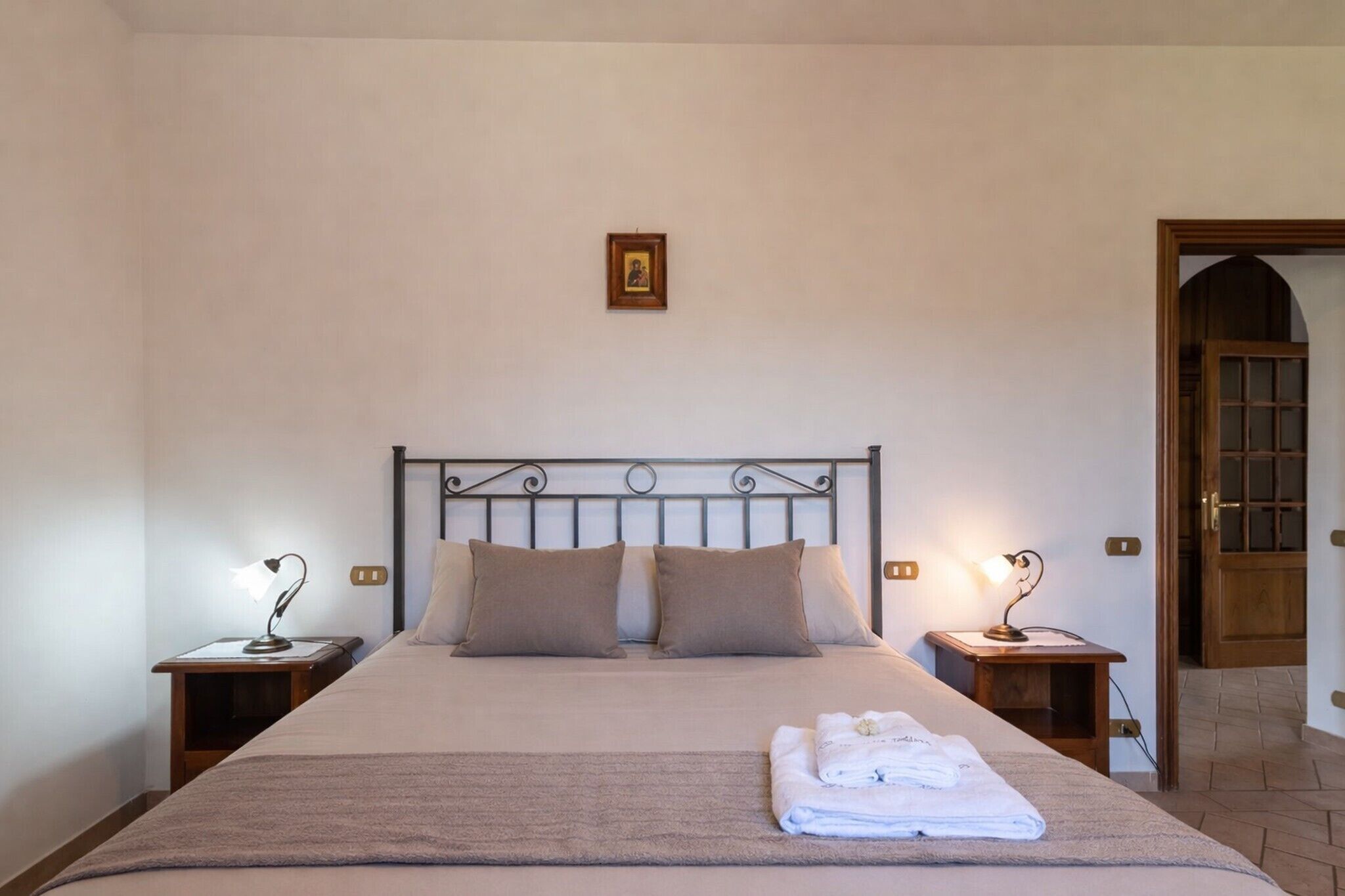 Ein ordentlich bezogenes Bett mit Kopfteil, zwei Nachttische mit Lampen, ein gerahmtes Bild an der Wand und eine Holztür zu einem anderen Raum.