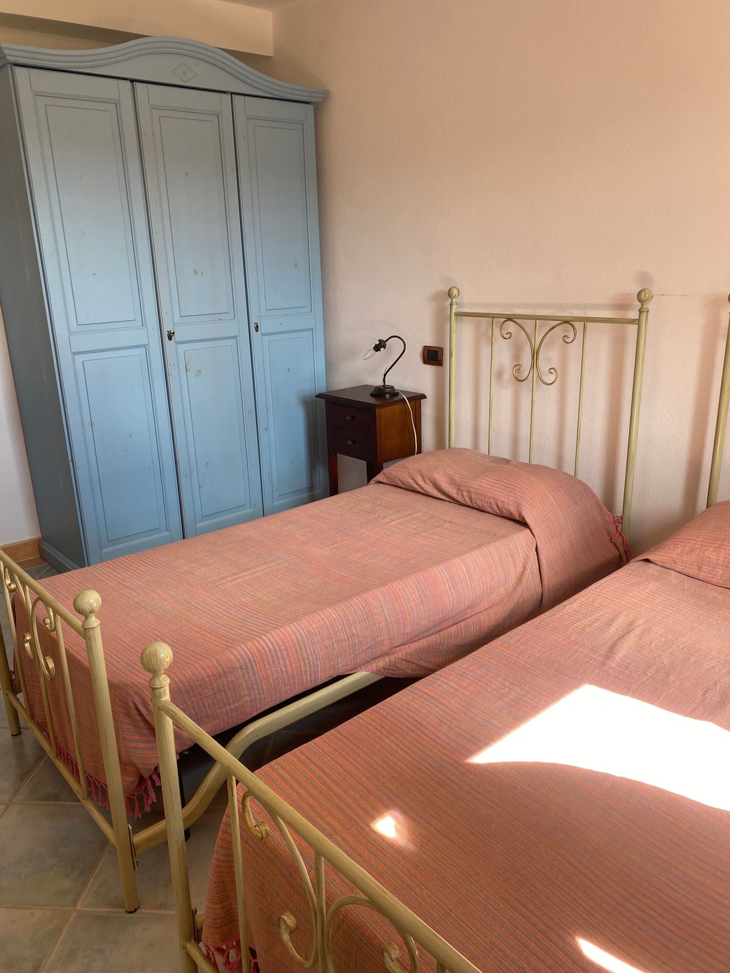 Une chambre avec deux lits simples, une armoire, une table de chevet avec une lampe et une tête de lit en métal.