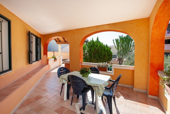A covered patio with a table and chairs, a potted plant, and a view of the sky and trees.