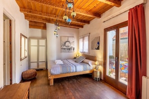 A bedroom with a wooden ceiling, a bed with patterned bedding, a hanging light fixture, and a view through a glass door.