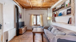 A cozy living room with a wooden ceiling, a sofa, a TV on a wooden stand, and a bookshelf with books and decorative items. - Holiday Home 'La Lechuga Azul' with Mountain View, Private Terrace and Wi-Fi (Guejar Sierra)