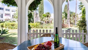 Une table avec une bouteille, un verre à vin et un plateau de fruits, placée dans un espace extérieur abrité, agrémenté d’arches et offrant une vue sur la verdure et les bâtiments.