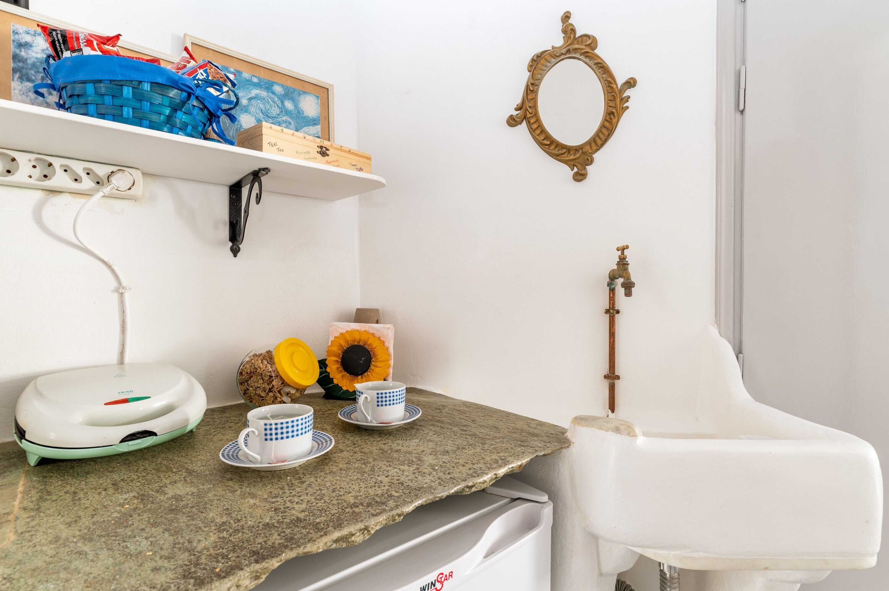 A kitchen with a countertop, a coffee maker, a sink, a mirror, and a shelf with various items.