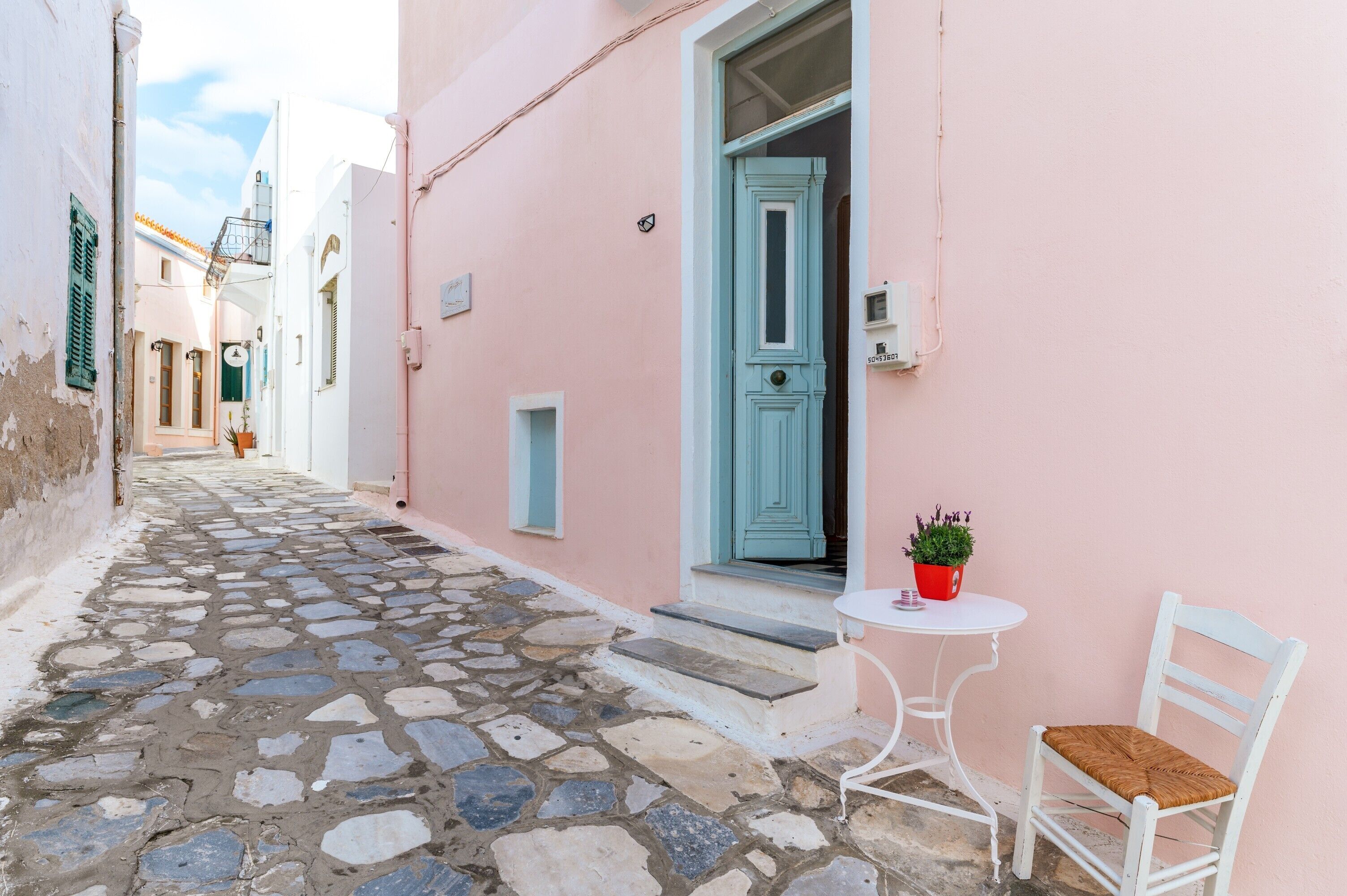 A narrow cobblestone street with a pink building, a blue door, and a white chair with a red pot of flowers.